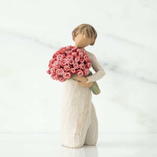 Small faceless girl figurine in white dress holding large pink roses boquet
